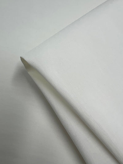 A close-up view of a folded piece of Super Cheap Fabrics Stretch Bengaline - Ivory - 150cm. The fabric appears smooth and soft, with clean, crisp edges visible against a light background. The image highlights the material's texture and folds.