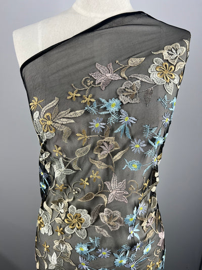 A dress form displays a sheer black mesh fabric adorned with intricate floral embroidery in pastel colors. The flowers are detailed in shades of blue, yellow, pink, and white, creating a delicate and elegant polyester pattern. The fabric drapes elegantly over the form with Embroidered Lace - Dolce - 150cm from Super Cheap Fabrics.