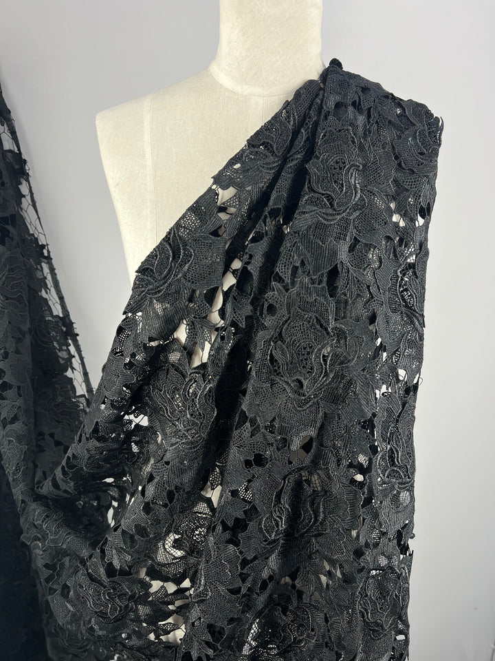 A mannequin is draped in Super Cheap Fabrics' Designer Anglaise Lace - Black - 120cm, intricately designed with a combination of sheerer and denser patterns. The 100% polyester fabric hangs elegantly over the shoulder and cascades down, showcasing its texture and detailed design against a plain white background.