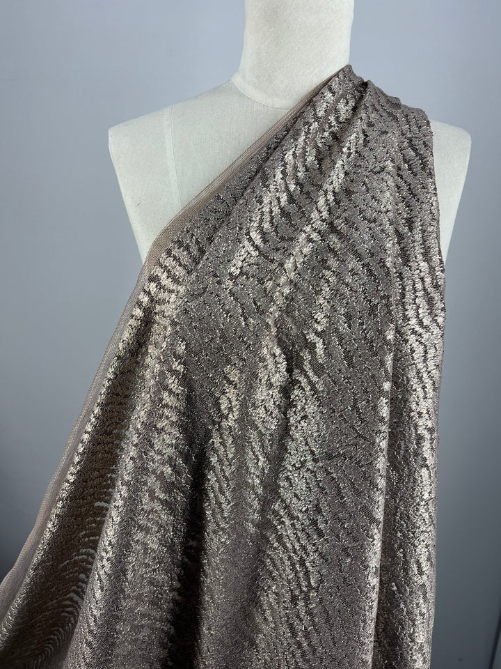 A mannequin draped with shimmering, textured fabric. The Evening Lace - Avalanche - 150cm by Super Cheap Fabrics has a subtle wavy pattern and a metallic sheen, with a mix of neutral tones. The background is plain and neutral, bringing focus to the intricate details of the sheer fabric.