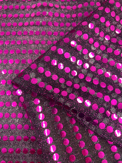 A close-up of Super Cheap Fabrics' Sequin - Baton Rouge - 112cm, displaying its sparkling 100% polyester fabric adorned with a pattern of shiny, metallic pink dots on a reflective dark background. The lightweight fabric is shown folded, highlighting its glossy and textured surface for a visually striking and vibrant look perfect for Baton Rouge’s lively scene.