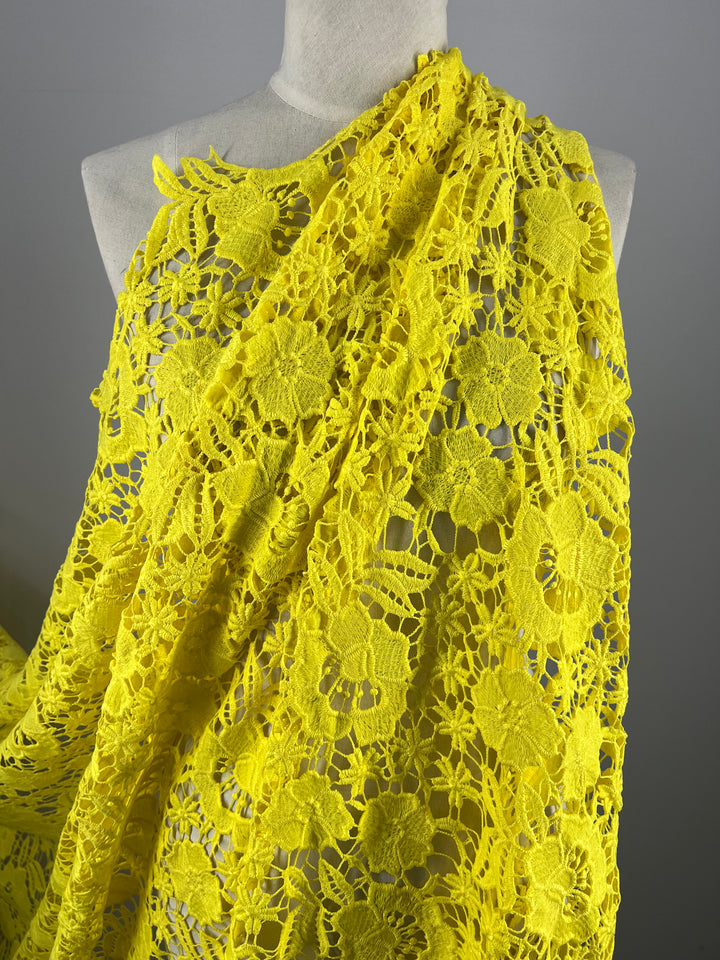 A mannequin draped in the vibrant Anglaise Lace - Yellow - 127cm from Super Cheap Fabrics, featuring intricate floral patterns, stands as an exquisite piece of home décor. The detailed openwork design showcases a stunning array of large and small flowers, creating an eye-catching and elegant look against the plain backdrop.