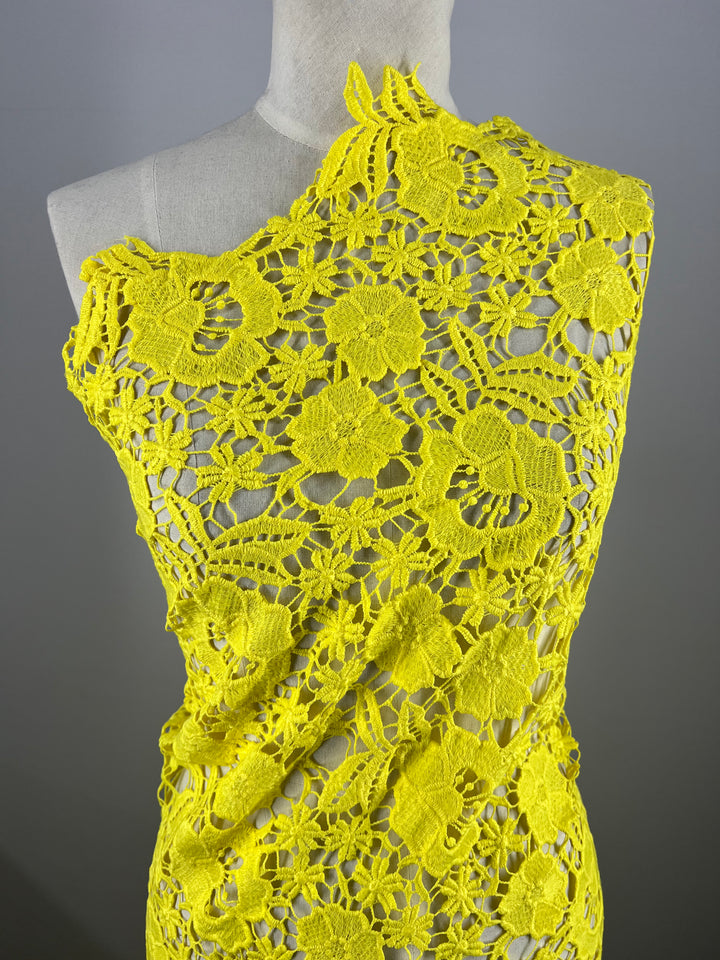 A mannequin showcases a vibrant yellow, intricately crocheted one-shoulder dress made from Super Cheap Fabrics' Anglaise Lace (127cm), adorned with large floral patterns. The asymmetrical design exposes one shoulder and highlights detailed craftsmanship akin to fine lace fabrics, set against a neutral gray background.