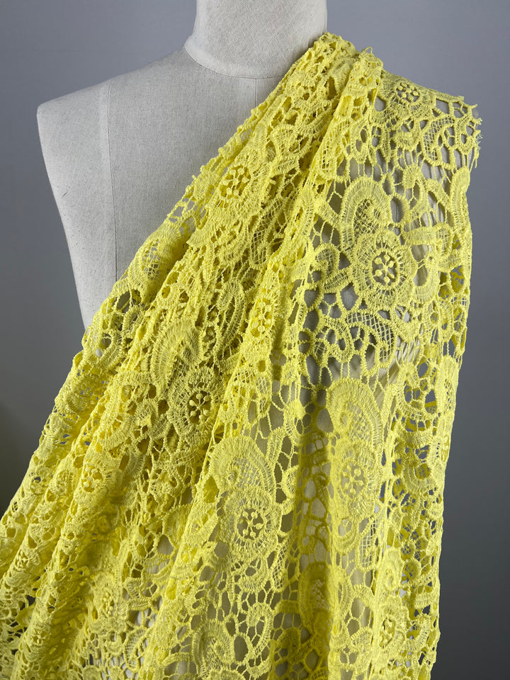 A mannequin draped in the Anglaise Lace - Limelight - 120cm from Super Cheap Fabrics showcases a vibrant yellow, lightweight lace fabric made of 100% polyester, featuring intricate floral and geometric patterns. The texture and delicate details of the lace are clearly visible, creating a visually appealing design against a plain, neutral gray background.