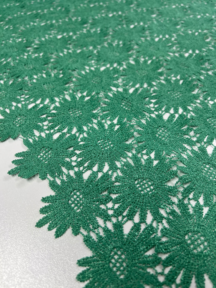 Close-up of the Anglaise Lace in Stone Green - 110cm from Super Cheap Fabrics, showcasing its intricate floral pattern. The detailed lace features flowers with centers resembling a delicate mesh structure, set against a white background. Perfect for bridal wear or as an elegant touch in home décor, the texture and craftsmanship are clearly visible.