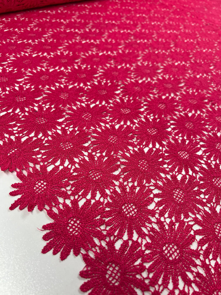 A close-up of the vibrant Hot Pink Anglaise Lace fabric from Super Cheap Fabrics, spread out on a flat surface. The 110cm lace pattern, often used in bridal wear, features detailed, interconnected flower designs with small circular centers, creating an intricate and delicate appearance perfect for home décor.