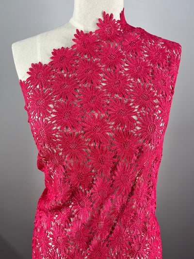 A mannequin draped in Super Cheap Fabrics' Anglaise Lace in Hot Pink, featuring intricate floral patterns. The lace fabric is adorned with detailed flower designs, creating a delicate and elegant appearance. The plain gray background highlights the texture and color of the 110cm wide lace.