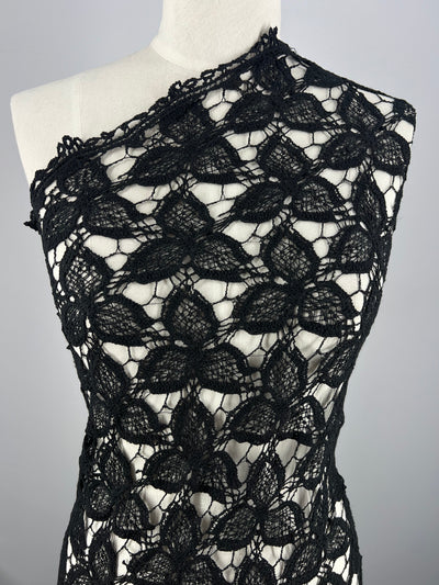 A mannequin is draped with Anglaise Lace - Black - 110cm from Super Cheap Fabrics, featuring a large floral pattern perfect for bridal wear. The lace is arranged asymmetrically across the mannequin's shoulder, showcasing the intricate detailing and delicate texture of the material against a plain gray background.