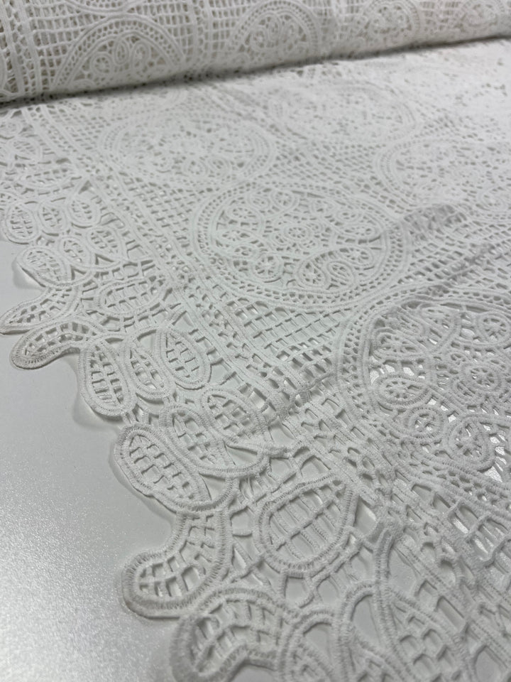 A close-up view of the intricate Anglaise Lace - White - 115cm by Super Cheap Fabrics highlights its detailed floral and geometric patterns. Perfect for bridal wear, this delicately woven lace features a scalloped edge that enhances its elegant appearance. Laid out on a smooth surface, the fabric showcases superior craftsmanship and is also suitable for home décor projects.