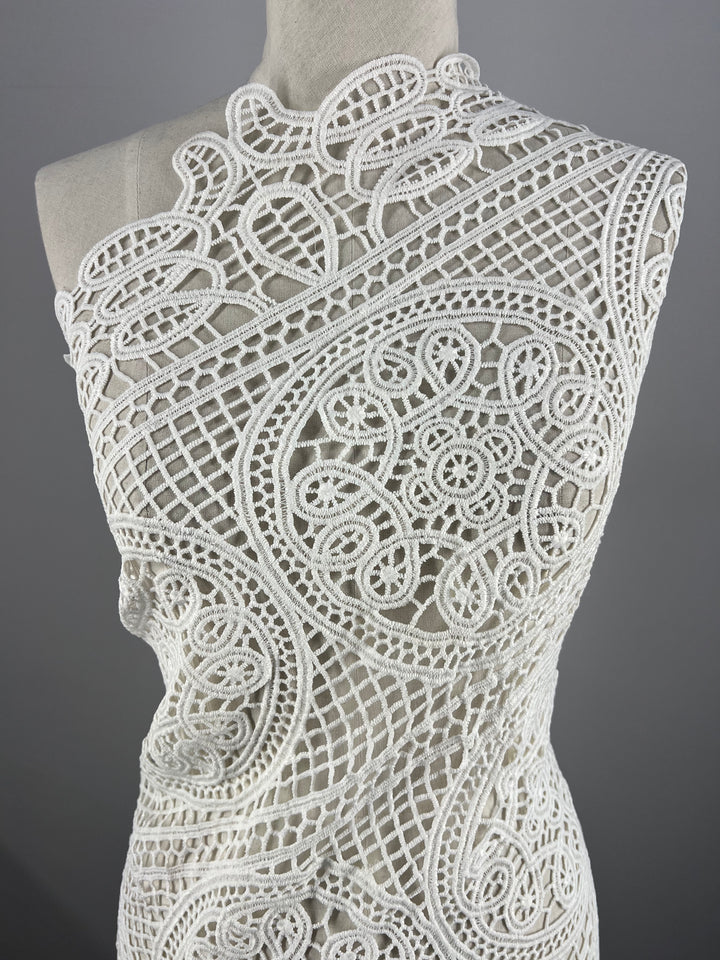 A close-up image showcasing the intricate detailing of the Anglaise Lace - White - 115cm by Super Cheap Fabrics, delicately draped over a mannequin. This elegant lacework, commonly used in bridal wear, features elaborate swirling and geometric designs that create a sophisticated texture against the neutral background.