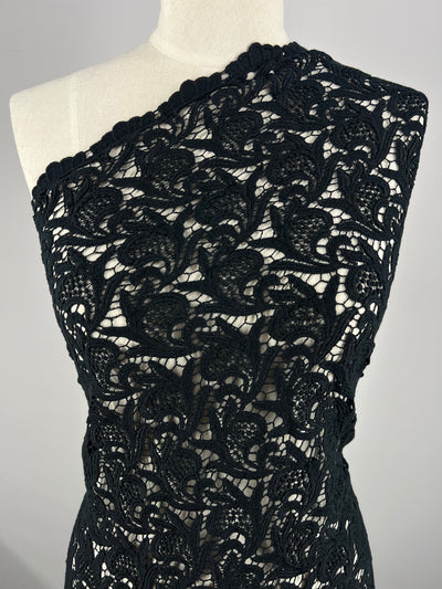 A mannequin is adorned with an elegant, single-shoulder garment crafted from the intricate "Anglaise Lace - GOTS Cotton - Black - 85cm" by Super Cheap Fabrics, featuring delicate floral and leaf patterns that add texture and detail against a plain, light-colored background. This exquisite piece exemplifies the sophistication found in formal wear.