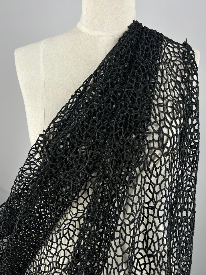 A white mannequin showcases the Anglaise Lace - Black - 125cm from Super Cheap Fabrics, elegantly draped over its shoulder. This partially transparent, intricately woven textile features a detailed pattern of interconnected loops and circles, making it ideal for home décor enthusiasts or those interested in bridal wear.