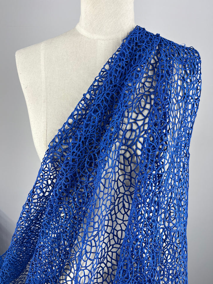 A close-up of a mannequin draped with a piece of intricate Anglaise Lace in Cobalt from Super Cheap Fabrics. This lace fabric, measuring 125cm, features a delicate, web-like pattern that showcases its detailed and open design. Perfect for bridal wear or home décor, the background is plain light gray, highlighting the vivid color and texture.