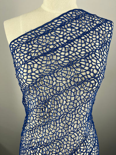 A mannequin is elegantly dressed in a one-shoulder creation made from Super Cheap Fabrics' Anglaise Lace - Cobalt - 125cm, featuring an intricate, open, circular pattern. The cobalt lace fabric drapes diagonally across the torso, showcasing the white base of the mannequin underneath.