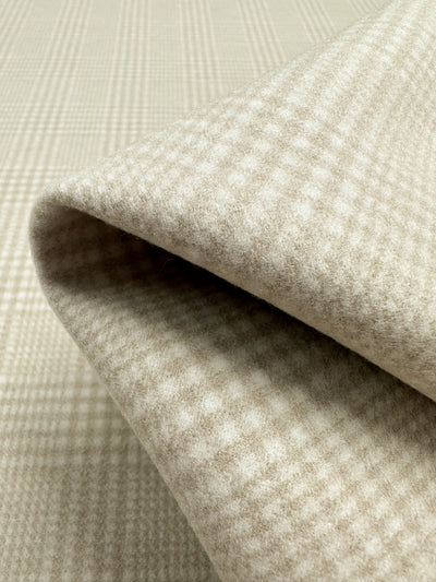 Folded beige Wool Flannel - Parchment - 148cm from Super Cheap Fabrics with a subtle plaid pattern placed on a matching fabric backdrop. The heavyweight fabric appears soft and woolen, ideal for coats and jackets, with fine vertical and horizontal lines creating a grid-like check design.