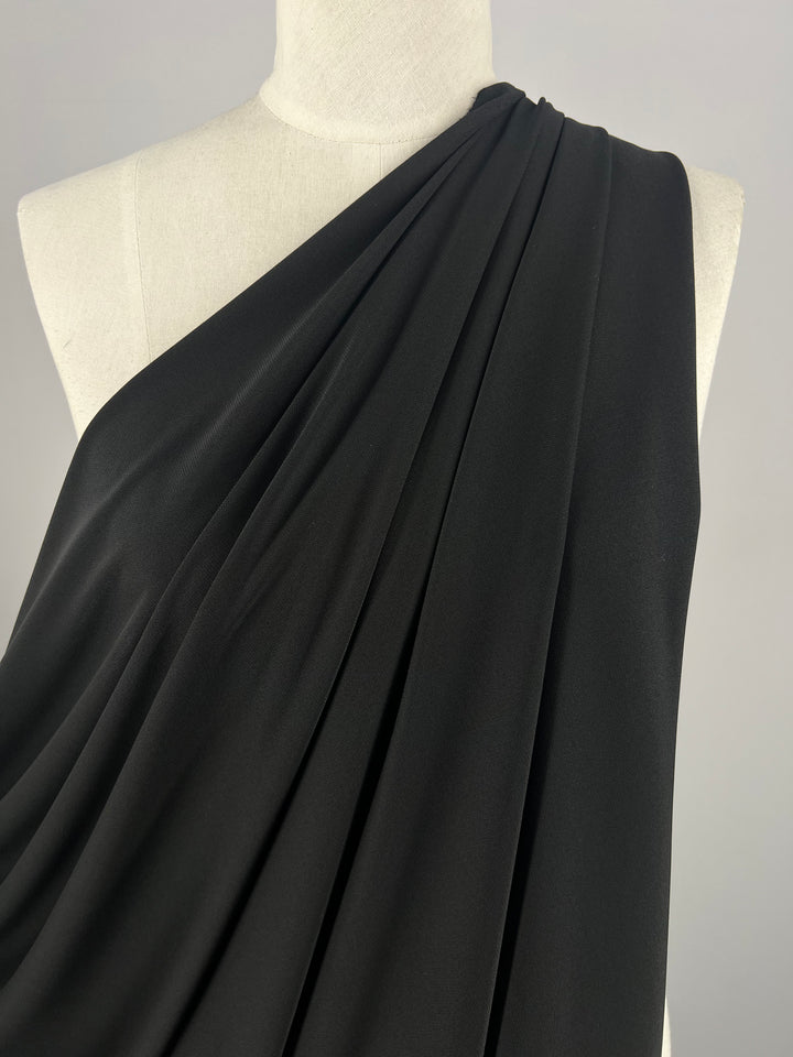 A close-up of a dress form showcases the "Double Knit Jersey - Black- 150cm" from Super Cheap Fabrics. The medium-weight fabric is artfully gathered, flowing elegantly over one shoulder, creating graceful folds against a neutral backdrop.