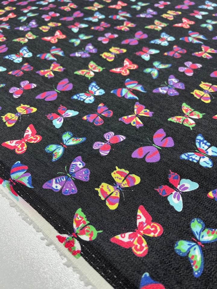 The Stretch Printed Denim Butterfly fabric by Super Cheap Fabrics is 145cm wide and features vibrant pink, blue, green, and yellow butterflies on a black background. This medium-weight material offers a lively design ideal for various projects.