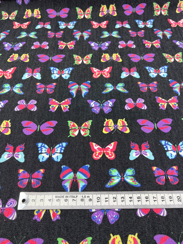 The Stretch Printed Denim Butterfly by Super Cheap Fabrics is a dark medium-weight fabric featuring colorful pink, blue, yellow, and green butterfly designs. Made from multi-use cotton, it includes a measuring tape at the bottom with "MADE IN ITALY" in centimeters. Width: 145cm.