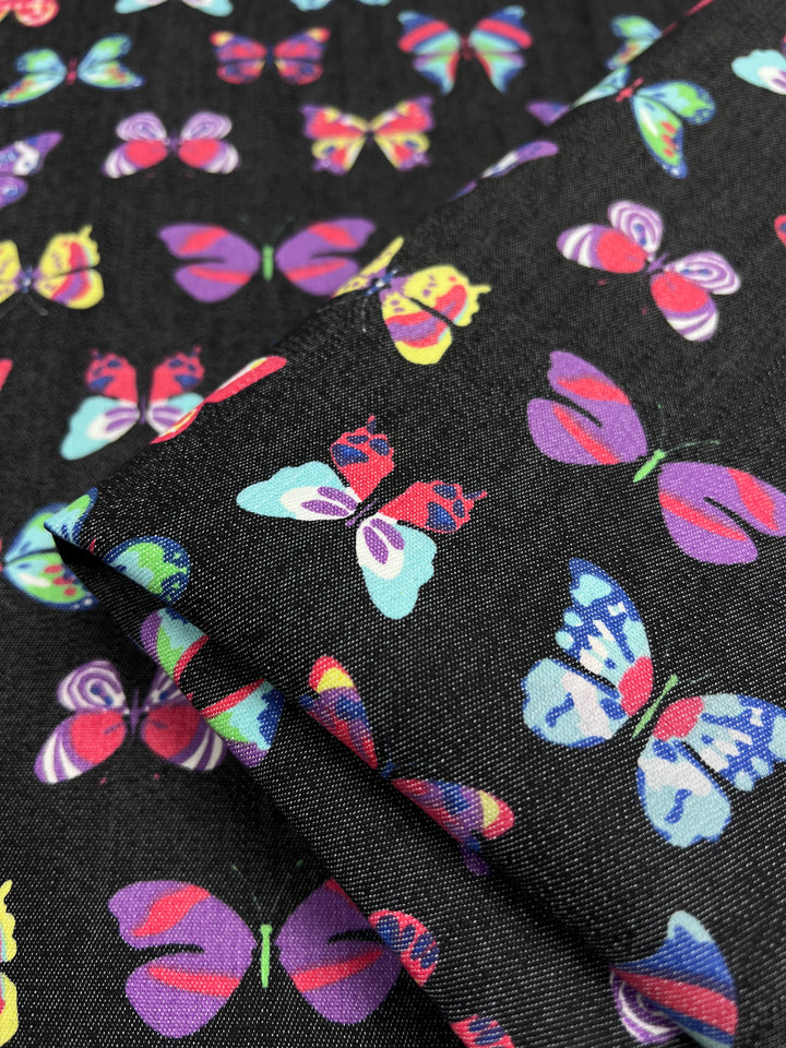 The Stretch Printed Denim Butterfly from Super Cheap Fabrics features vibrant butterflies in purple, red, blue, and yellow on black cotton. Its medium-weight texture is perfect for versatile projects, with folds that highlight the stunning butterfly patterns. Width: 145cm.