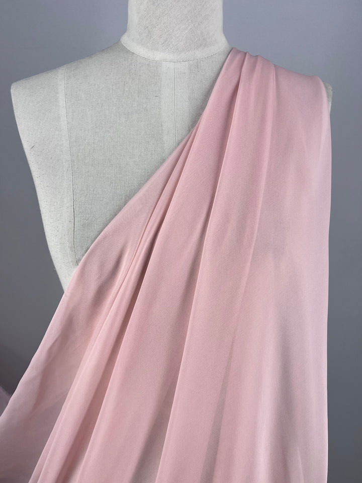 A mannequin elegantly displays Super Cheap Fabrics' Pure Silk Georgette - Pink Lotus - 135cm, draped like a toga with the lightweight fabric covering one shoulder, creating gentle folds that contrast beautifully with the neutral gray background.