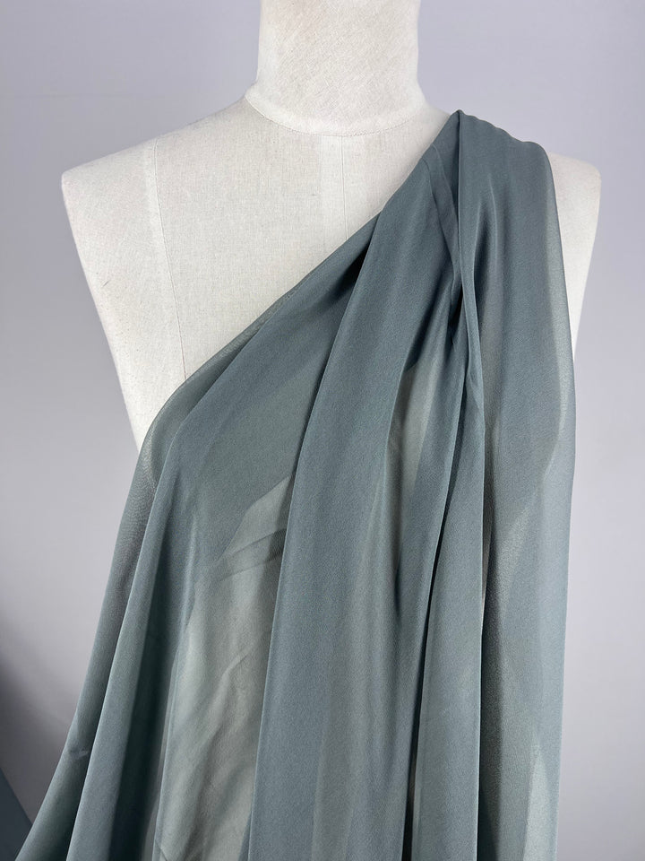 Displayed on a mannequin is the Super Cheap Fabrics brand's Pure Silk Georgette in Mercury, measuring 135cm. This semi-transparent, dark gray fabric drapes diagonally over one shoulder, flowing down the torso for elegant spring outfits against a plain light gray backdrop.