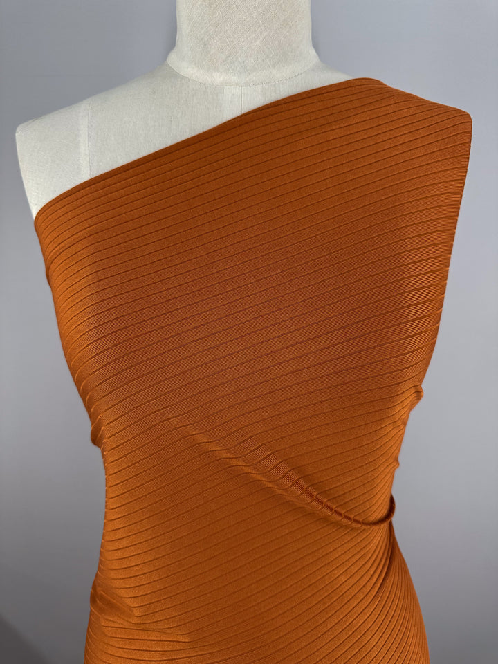 A mannequin models the "Rib Knit - Rust - 175cm" dress by Super Cheap Fabrics, crafted from medium weight rib knit with diagonal lines and a fitted silhouette. The rust-colored, one-shoulder dress with generous stretch stands out against a plain gray background.