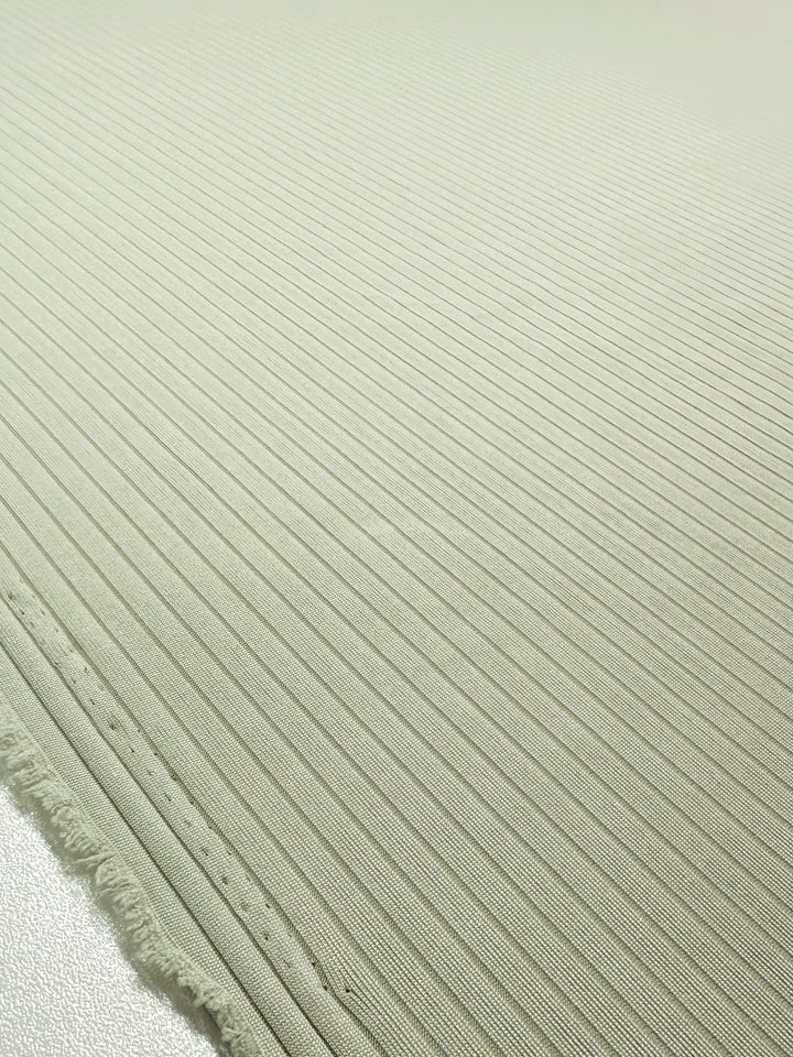 Close-up of Super Cheap Fabrics' Rib Knit in Celadon Tint, 168cm wide, showcasing thin parallel ridges on a light beige, medium weight fabric. The frayed bottom edge adds a casual touch to its textured look.