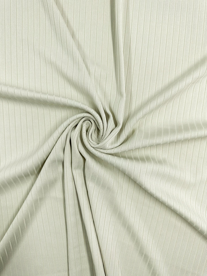 The Rib Knit - Celadon Tint by Super Cheap Fabrics features a close-up of pale green ribbed fabric gathered into a central swirl. This stretchy, medium-weight knit shows visible vertical lines for a subtle texture and pattern. Width: 168cm.