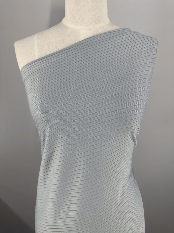 A mannequin displays a light pearl blue, ribbed, one-shoulder dress crafted from Rib Knit fabric by Super Cheap Fabrics. This medium-weight, super stretchy material with horizontal ribbing hugs the mannequin's form against a neutral gray background.