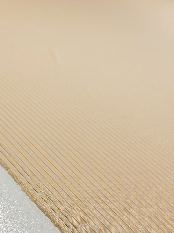 Close-up of the "Rib Knit - Apricot Gelato - 170cm" fabric by Super Cheap Fabrics, displaying a light beige, medium weight rib knit with thin parallel lines. The frayed edge is visible on the left against a softly out-of-focus background in a similar tone.