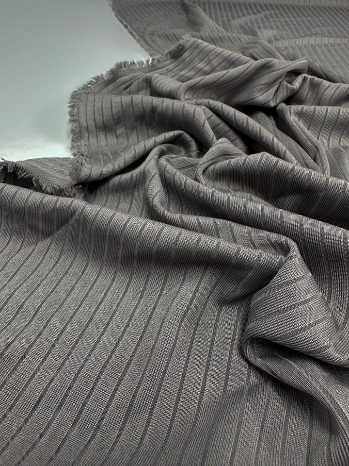 Super Cheap Fabrics' Rib Knit in Dark Grey (170cm) is draped on a light surface. This medium-weight fabric's vertical ridges add texture and depth, frayed edges appear on one side, and the lighting highlights its stretchy, comfortable nature.