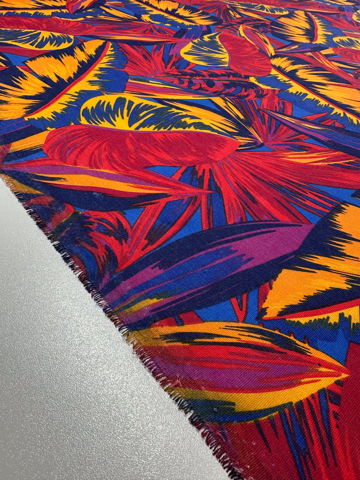 A close-up of Super Cheap Fabrics' Cotton Duck - Palm Scarlet - 140cm reveals its abstract tropical pattern in vivid red, yellow, orange, and blue. The slightly frayed edge on a smooth gray surface evokes a Flame Scarlet hue—ideal for striking home décor.