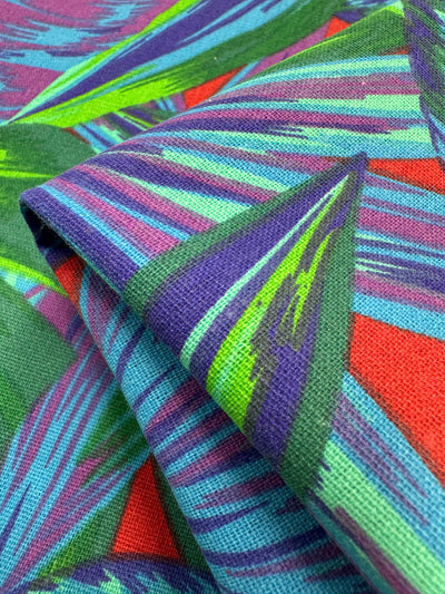 This Cotton Duck fabric by Super Cheap Fabrics, called Palm Lush Meadow, features vibrant streaks of green, blue, purple, and red on a textured medium-weight 140cm textile. With a folded corner enhancing its dynamic pattern, it adds energy and movement to any home décor.