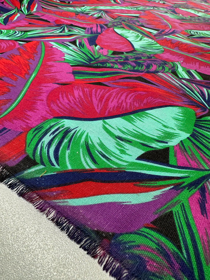 Elevate your home décor with Super Cheap Fabrics' "Cotton Duck - Palm Cockatoo." This 140cm medium-weight cotton fabric showcases vibrant green, pink, and red tropical leaf patterns on a textured surface with visible fringe for a striking look.