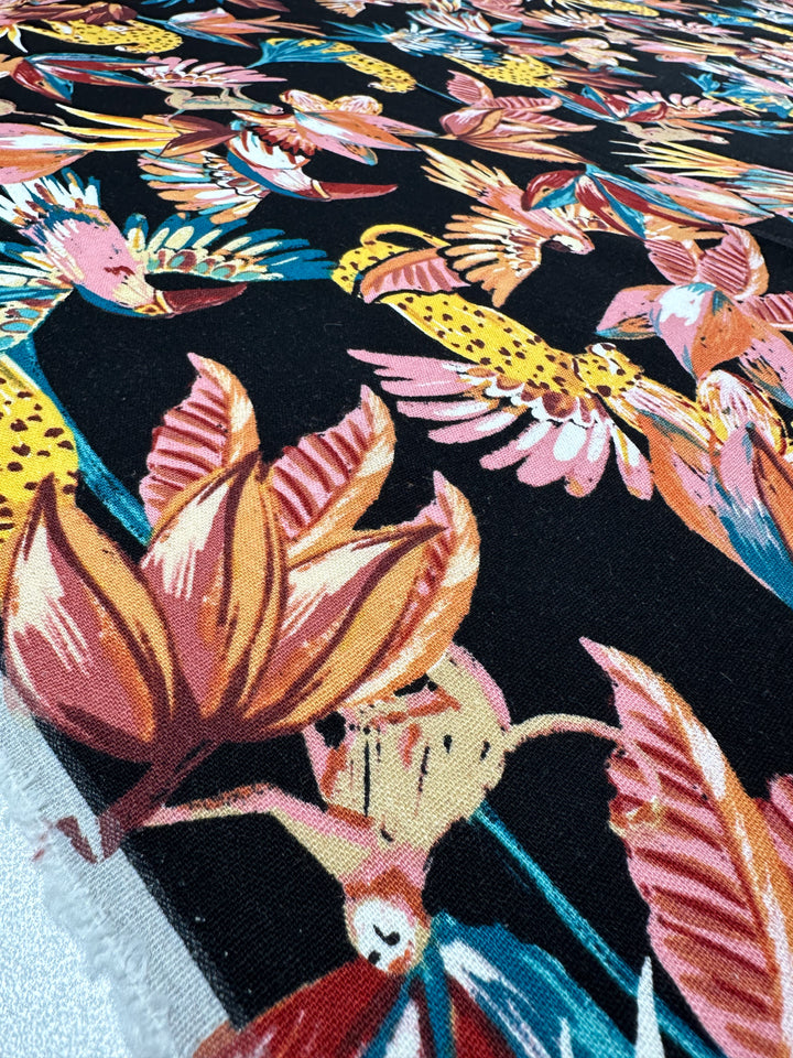 A close-up of the lightweight Linen Blend - Paradiso Black fabric by Super Cheap Fabrics highlights a vibrant tropical pattern featuring birds and flowers in pink, orange, and yellow on a black background, creating a lively and colorful look. Width: 135cm.