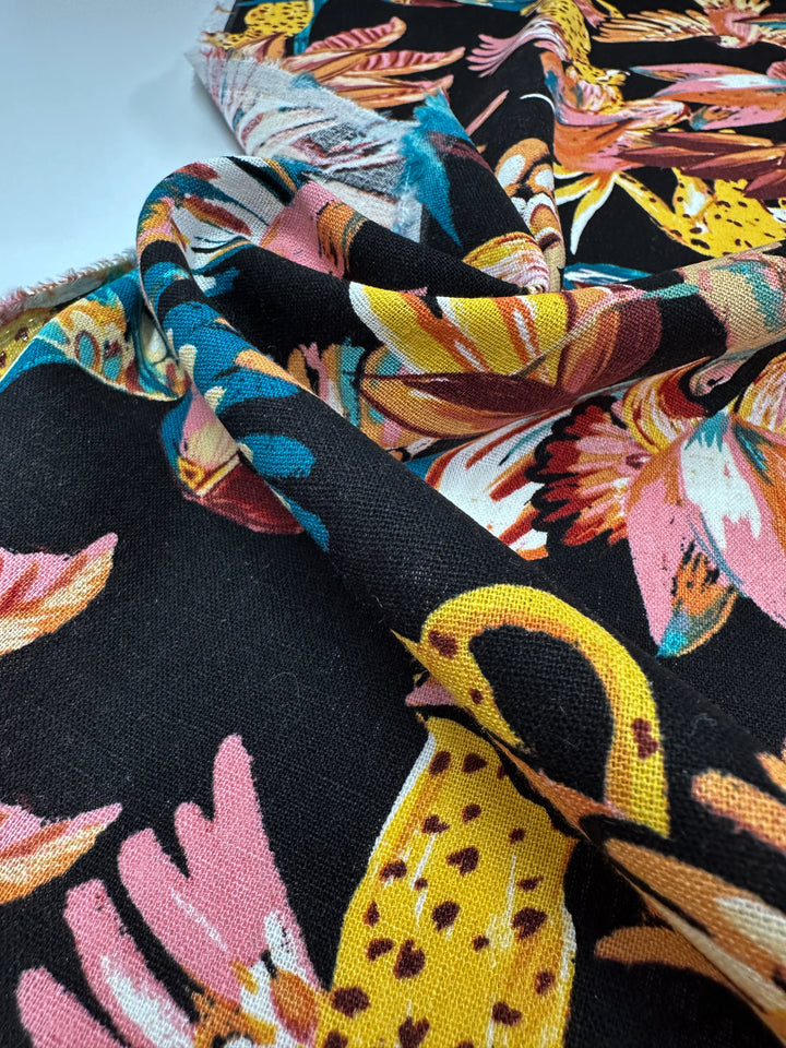 A close-up of the Super Cheap Fabrics' Linen Blend - Paradiso Black 135cm shows a vibrant leaf and abstract pattern in pink, yellow, blue, and white. The lightweight fabric is elegantly draped to showcase its texture and flow.