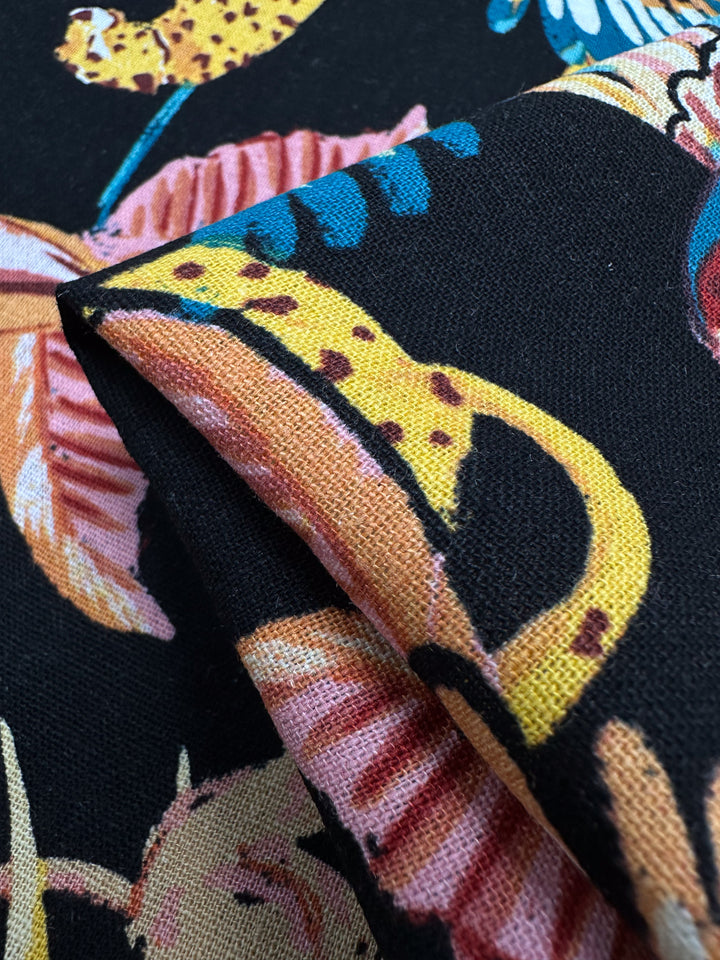 Close-up of Super Cheap Fabrics' Linen Blend - Paradiso Black. This vibrant tropical-themed fabric, 135cm wide, features leopards, leaves, and flowers in bold detail on a black background; it is lightweight and neatly folded to showcase its intricate design.