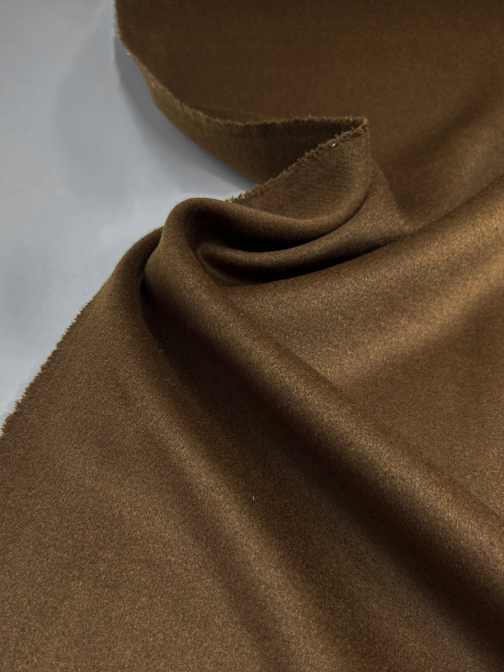 Close-up of Super Cheap Fabrics' "Wool Cashmere - Emperador - 150cm," showcasing its smooth, soft brown fabric with a slight sheen and gentle folds on a white surface. The premium material's contours are elegantly highlighted by subtle shadows.