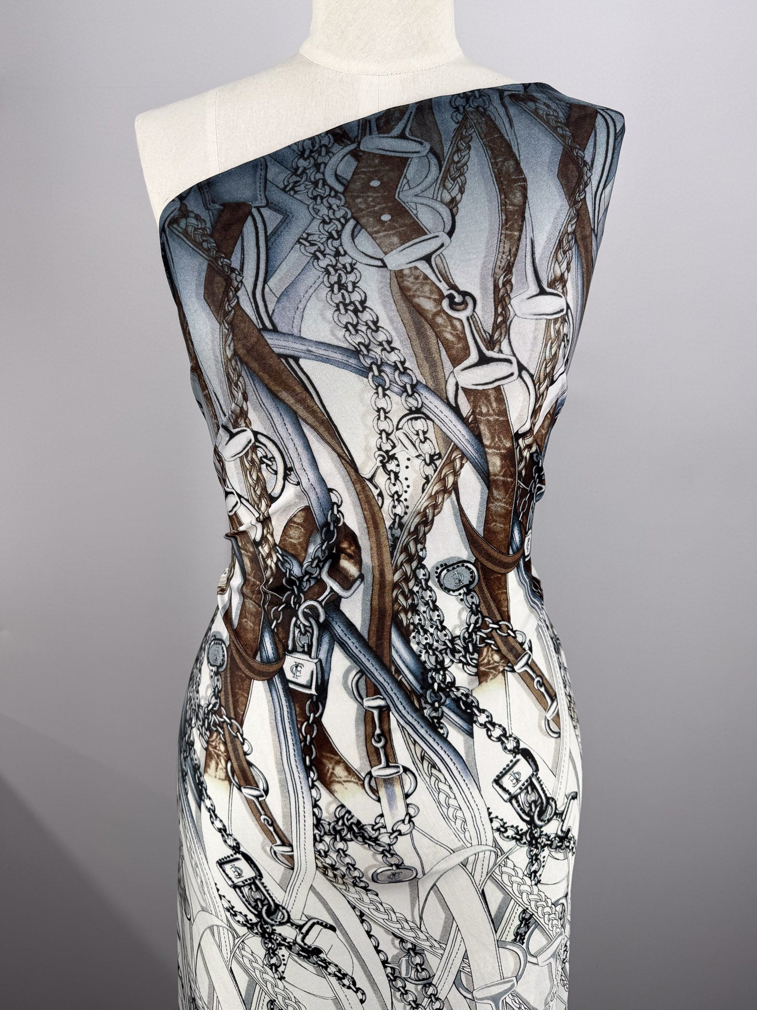 A mannequin displays Super Cheap Fabrics' Viscose Knit - Link (138cm), a stretch fabric with an elegant pattern of brown leather straps, silver chains, and buckles over a white and gray background.