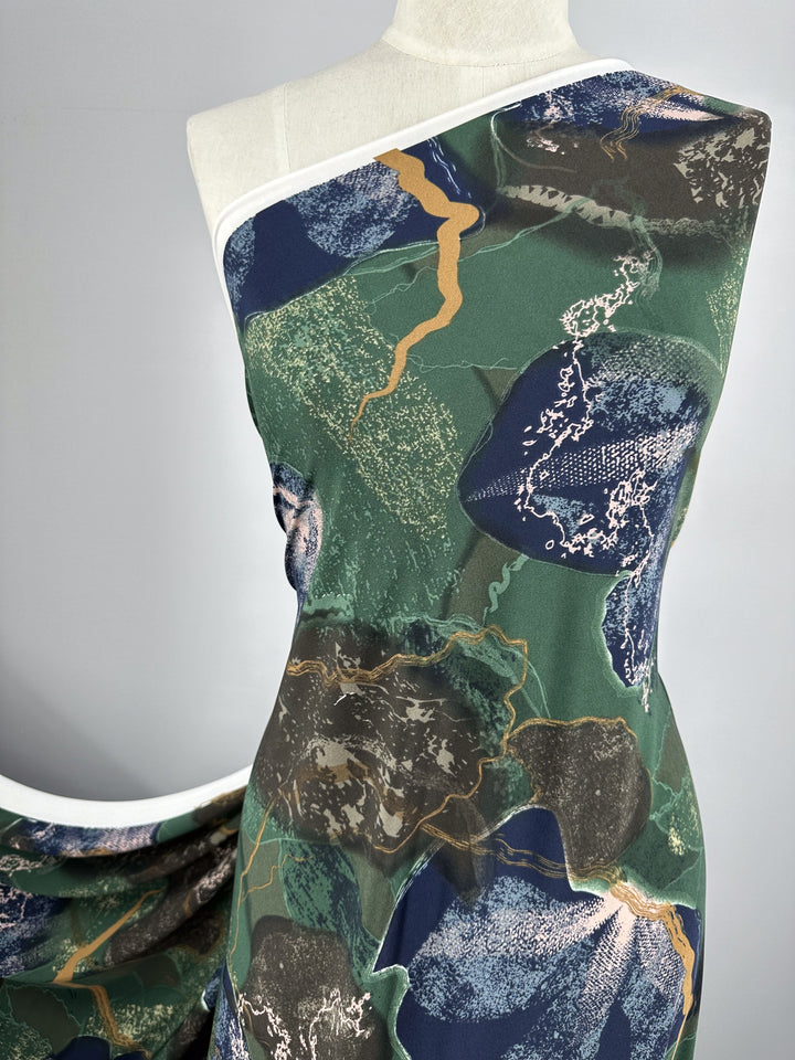 A mannequin is draped with a single-shoulder garment featuring a modern abstract pattern. The medium-weight fabric design includes shades of green, blue, gold, and white with organic, irregular shapes and textured details. The background is a plain gray. The fabric used is the Printed Lycra - Drifter - 150cm from Super Cheap Fabrics.