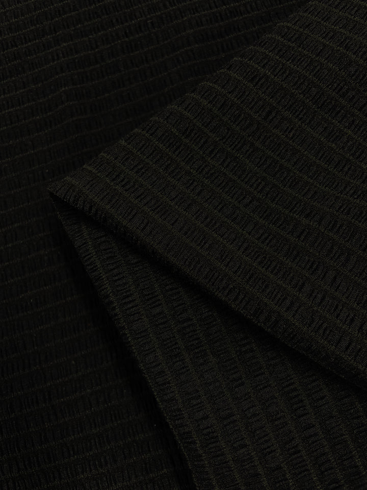 A close-up of the Designer Mesh - Black Rouge from Super Cheap Fabrics showcases its textured black fabric with a ribbed pattern. The 130cm wide, lightweight polyester spandex material appears to be folded, creating a layered effect. The fabric has a subtle sheen, and the vertical ribbing adds dimension to the overall look.