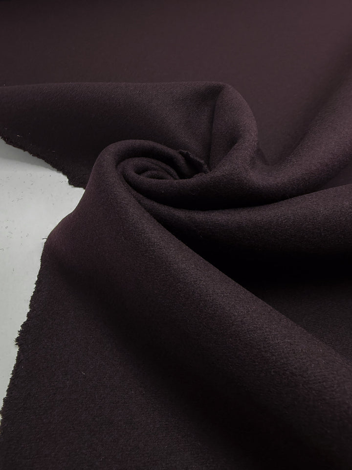 A close-up of Wool Cashmere - Gothic Grape by Super Cheap Fabrics, 150cm wide, featuring dark purple fabric with luxurious softness, partially rolled in a swirl to display unfinished edges on a flat surface.