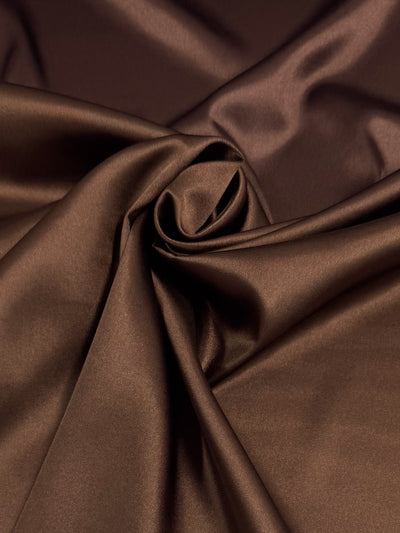 Satin Deluxe - Chocolate Lab by Super Cheap Fabrics is a smooth, glossy brown satin fabric (150cm wide), draped in loose folds and central spiral, creating soft highlights and shadows.