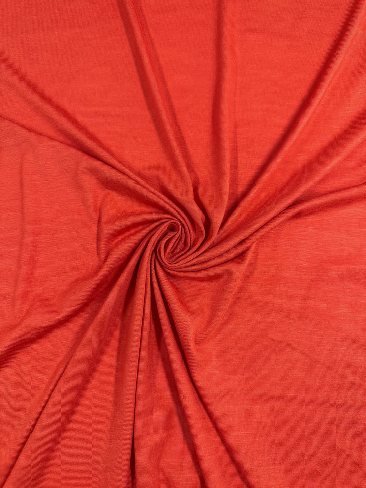 A close-up of a piece of Rayon Lycra - Hot Coral - 145cm from Super Cheap Fabrics arranged in a swirling pattern. The material appears smooth and has a faintly visible texture. The medium weight fabric radiates out from the center swirl, creating soft folds and varying shades of red.