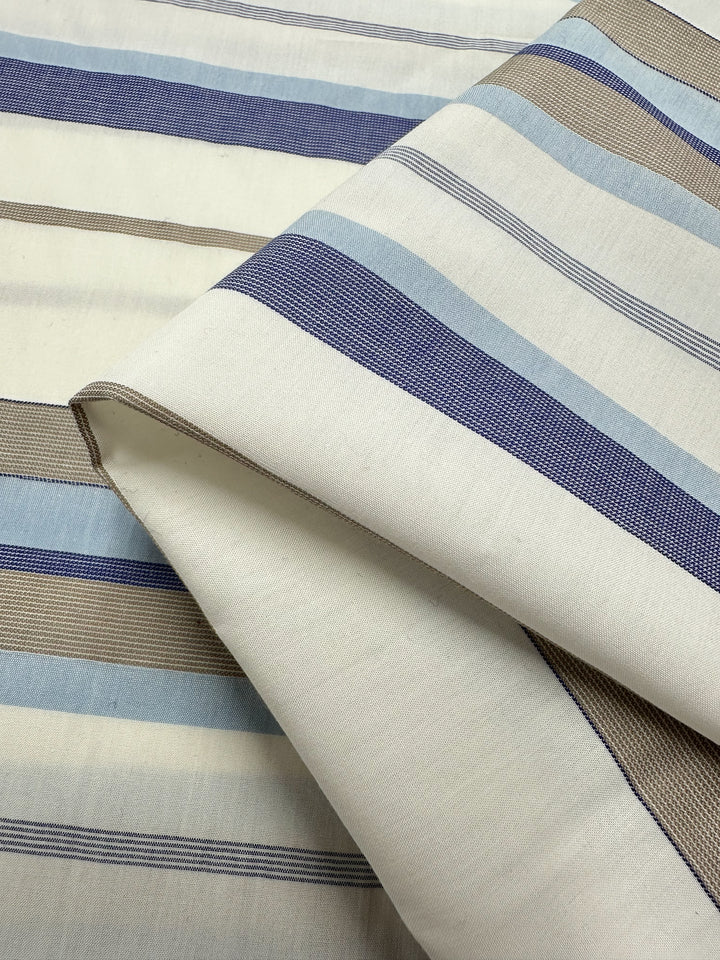 Close-up of a folded Super Cheap Fabrics' Cotton Voile - Blue Breeze - 155cm, showcasing horizontal stripes in varying widths. The stripes feature shades of blue, beige, and gray on a white background. Its smooth texture makes it ideal for light and airy garments, accentuating the alternating colors and stripe sizes.