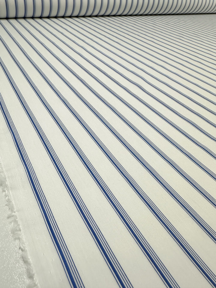 A close-up view of a roll of Super Cheap Fabrics' GOTS Cotton - Thin Regatta Stripe - 150cm, featuring thin blue vertical stripes. The perspective highlights the continuous length of this certified organic cotton fabric roll with the stripes running parallel from top to bottom in the image.