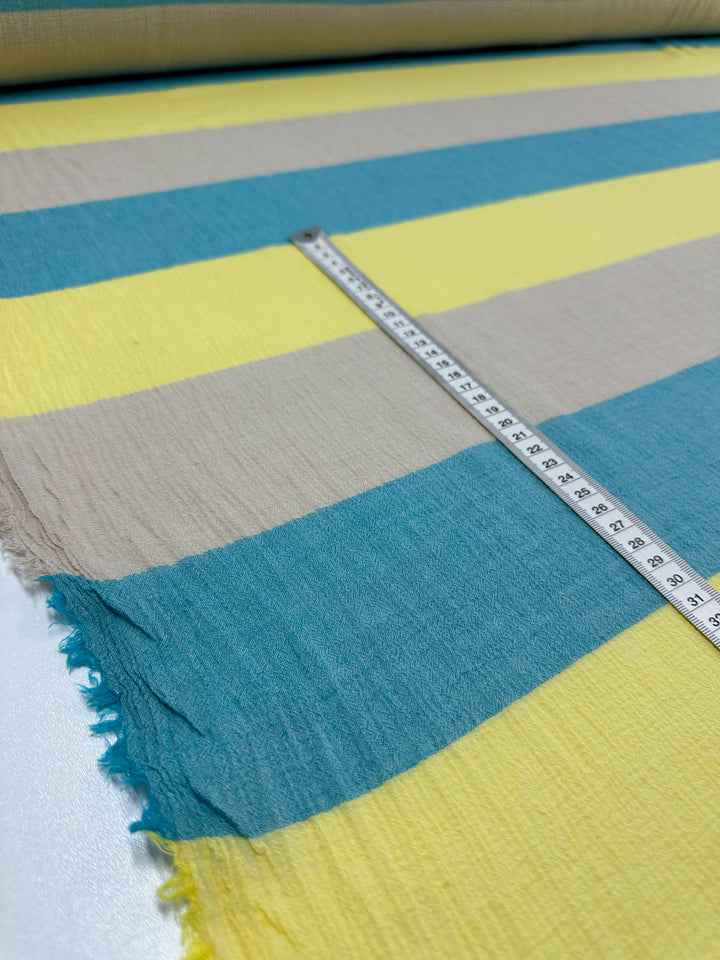 A close-up photograph features the lightweight "Cotton Gauze - Beachy - 130cm" by Super Cheap Fabrics. The fabric showcases wide horizontal stripes in blue, neon yellow, and beige. A measuring tape runs horizontally on top, displaying measurements in centimeters. The slightly frayed edges suggest it could be ideal for children's clothing or home decor projects.