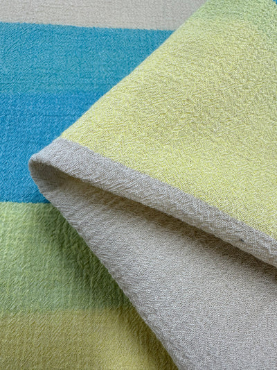 This close-up of Super Cheap Fabrics' Cotton Gauze - Beachy - 130cm showcases its lightweight material with blue and green horizontal stripes. A corner is folded over to reveal the light grey underside, highlighting the soft, slightly raised fibers that lend a tactile feel, making it ideal for children's clothing or home decor accents.