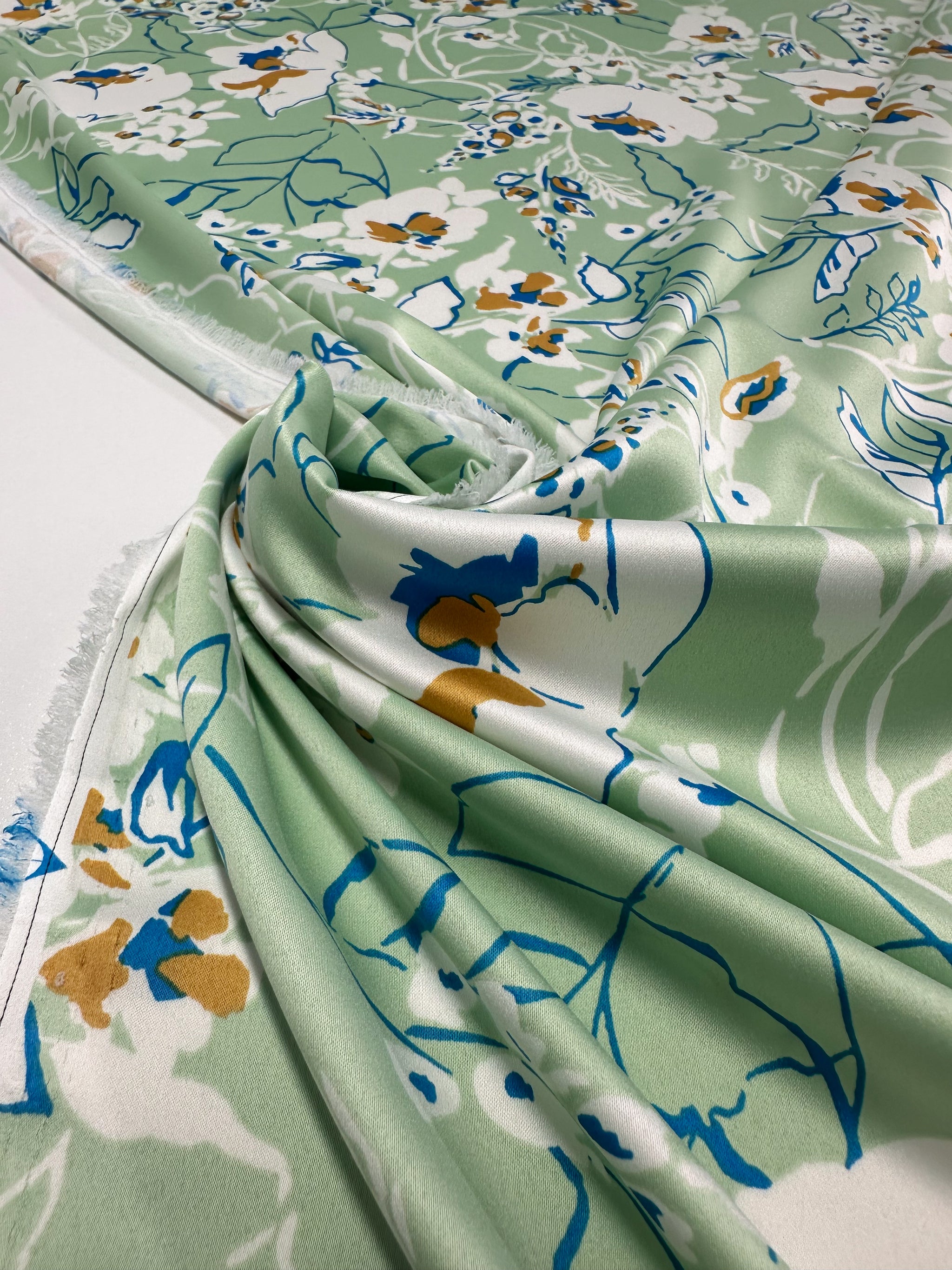 A close-up of elegant pieces in luxurious Printed Stretch Satin - Green Sakura by Super Cheap Fabrics, showcasing vibrant floral patterns in white, yellow, and blue. The fabric's gentle drape enhances its texture and design, with visible selvage on the left.
