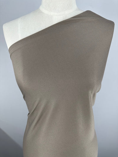 A mannequin draped in light brown, off-the-shoulder fabric on a neutral gray background. The lightweight 100% polyester material appears smooth and is slightly gathered at the sides. The stone color enhances the elegant simplicity, with the mannequin's head and arms not visible. This is **Interlock - Stone - 150cm** by **Super Cheap Fabrics**.